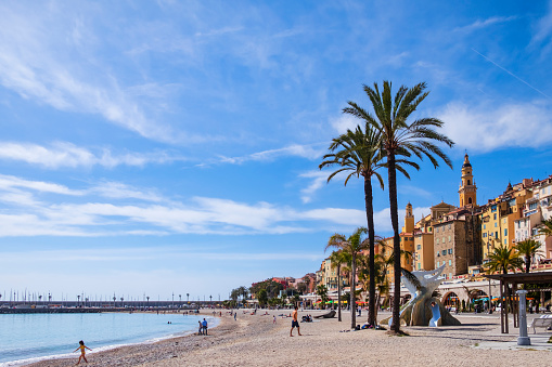People enjoying a warm sunny spring day on the Plage des Sablettes in Menton, whose promenade is bordered by the characteristic colorful buildings