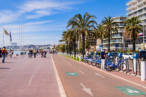 A typical sunny day on the Promenade des Anglais: while young men are renting a bike, others, tourists and locals, are strolling on this very popular promenade along the Mediterranean coast of Nice