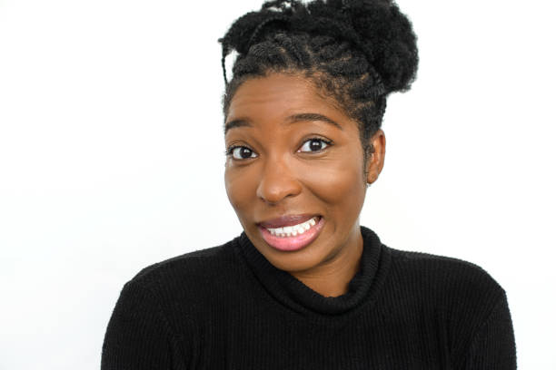 Uneasy African American woman in studio. Female model in turtleneck with curly hair looking at camera, smiling. Portrait, studio shot, emotion concept