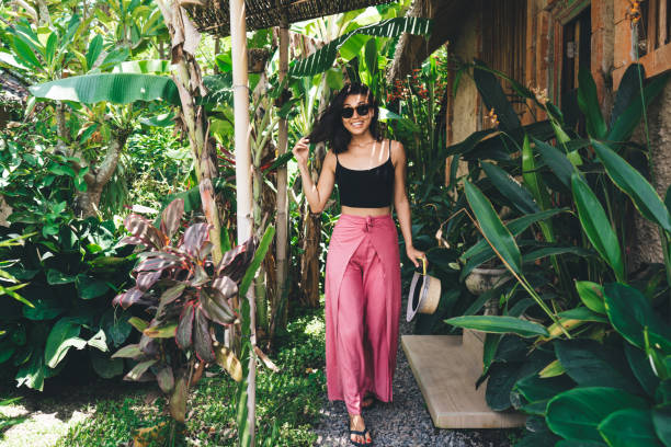 Delighted female traveler in colorful clothes smiling and looking at camera during stroll on wooden porch near blooming plants and trees