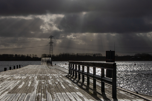 Landscape with stormy clouds and sunrays over a wooden pier with black birds facing the wind and a dark lake. In the distance, there are high poplar trees together with an electricity mast and cables. High-quality photo