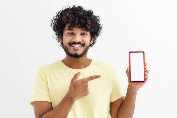 istock Happy Indian man student standing with mobile phone, pointing his finger at the empty screen on white background, looking at camera and smiling 1423121982