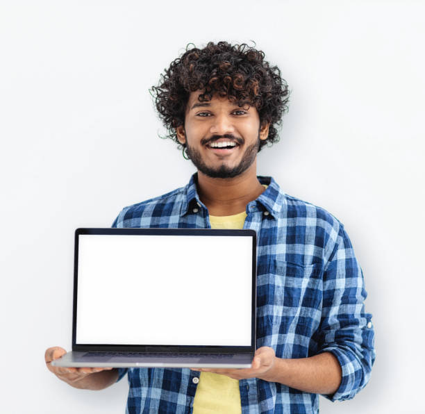istock Happy Indian man student standing with open laptop with blank white mockup screen on white background, looking at camera and smiling 1423122043