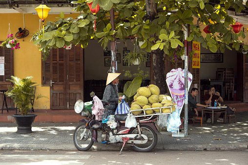 Hoi An, Vietnam - May 07, 2018: Old woman with traditional conical hat (Non La) selling durian fruit from her motorcycle on the street.