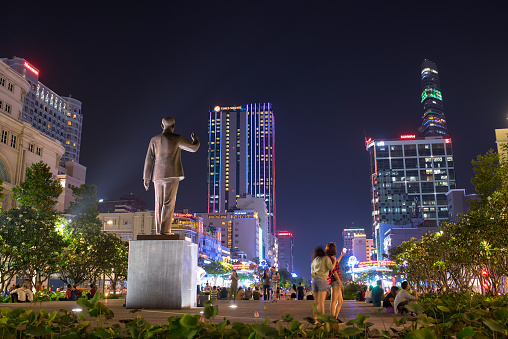 Ho Chi Minh City, Vietnam - May 03, 2018: New Ho Chi Minh statue and tourists taking photos with smartphone on Nguyen Hue Walking Street with illuminated modern buildings at night.