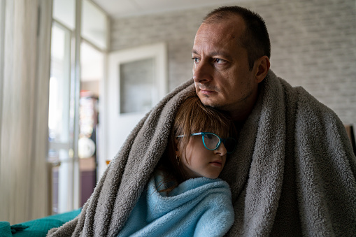 Dissatisfied father and daughter having problem with central heating, sitting on sofa at home, freezing. Freezing family warm blankets looking at camera while sitting on sofa in cold kitchen