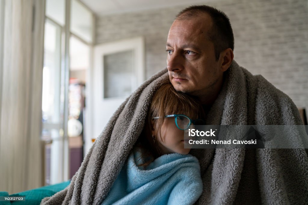 Cold at home Dissatisfied father and daughter having problem with central heating, sitting on sofa at home, freezing. Freezing family warm blankets looking at camera while sitting on sofa in cold kitchen Cold Temperature Stock Photo