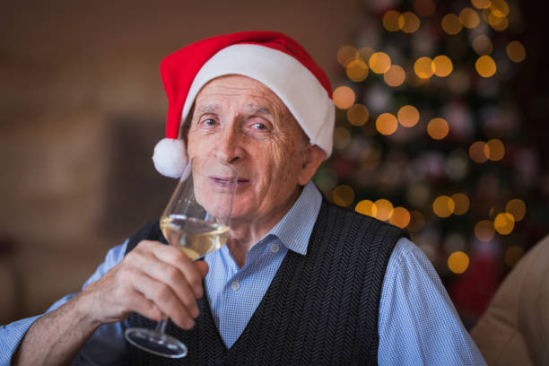 homem sênior feliz usando o chapéu do papai noel ao beber champanhe em casa - new years day new years eve new year ethnic - fotografias e filmes do acervo