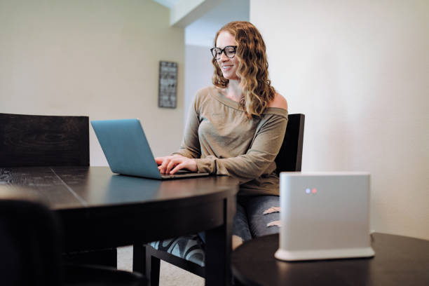 Woman Using Laptop in Apartment Dining Room A mid-adult Caucasian woman works at a laptop computer at a dining room table. A high speed network router visible in the foreground. router stock pictures, royalty-free photos & images