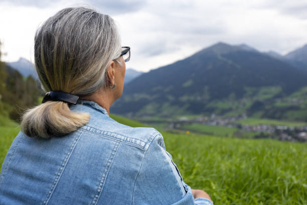 an elderly woman in a denim suit sits on a high meadow and looks at the mountains on the opposite side of the gorge. - footpath field nature contemplation imagens e fotografias de stock