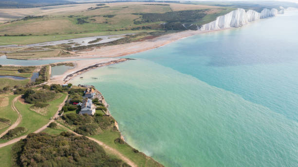 Aerial view of scenic footpaths, 19th-century cottages, flood plains where River Cuckmere meets English Channel, and famous chalk cliffs. East Sussex, summer.