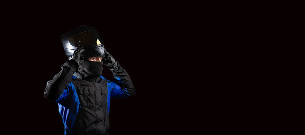 male motorcyclist in a motorcycle outfit and a balaclava puts on a helmet on a dark background. The concept of protective equipment for a biker.