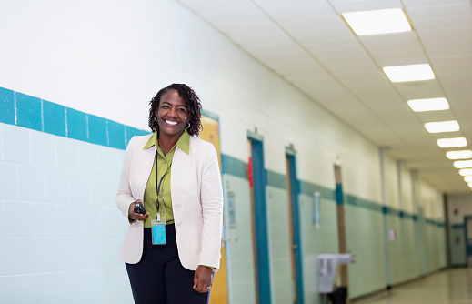 A mature African-American woman standing in a school hallway, looking at the camera, holding a walkie talkie. She is a teacher, school administrator or principal.