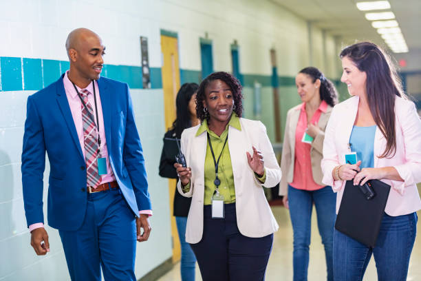 Multiracial group of teachers walking in school hallway A multiracial group of five teachers or school administrators conversing as they walk through a school corridor. The focus is on the three in the foreground. The African-American woman in the middle, in her 50s, is talking while her coworkers look at her and listen. school building stock pictures, royalty-free photos & images