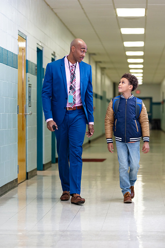 An elementary student walking with a teacher, or perhaps the school principal, through the school hallway. The student is an 8 year old multiracial boy, African-American and Caucasian. The teacher is a mature African-American man, in his 40s. They are side by side, looking at each other.