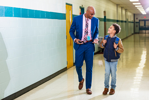 An elementary student walking with a teacher, or perhaps the school principal, through the school hallway. The student is an 8 year old multiracial boy, African-American and Caucasian. The teacher is a mature African-American man, in his 40s. They are side by side, looking at each other, smiling.