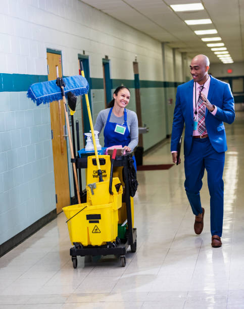 Teacher walks through school hallway, waves to custodian A mature multiracial woman in the hallway of a school working as a janitor pushing a cart of cleaning supplies. She is in her 40s, African-American and Caucasian. She smiles at an African-American man wearing a suit, walking by and waving to her. He is a teacher, or perhaps the school principal. custodian stock pictures, royalty-free photos & images