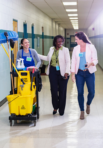 A multiracial group of three mature women walking through a school corridor. The mixed race woman on the left is a custodian pushing a cleaning cart. The African-American woman, in the middle, a teacher or school principal, greets the custodian with a pat on the back as she walks by, talking with another teacher.