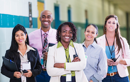 A multiracial group of five teachers or school administrators standing together in a school hallway smiling confidently at the camera in a group portrait. The focus is on the African-American woman in the middle. She is the team leader, perhaps the school principal. She is in her 50s and her coworkers are in their 40s.
