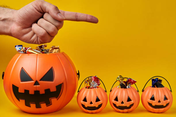istock Hand points away by big Halloween pumpkin head basket with three small baskets full of candy over yellow background with copyspace 1423170122