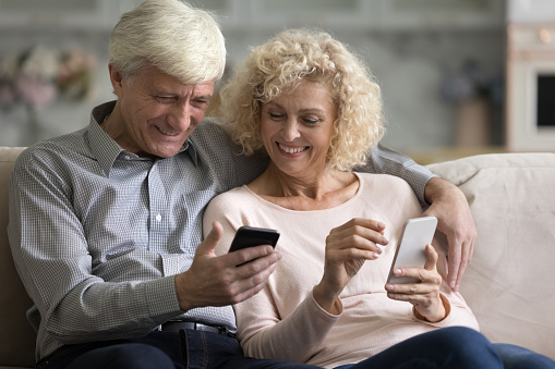 Joyful happy mature married couple using two mobile phones, learning to use online app together, sharing data, media content, shopping on Internet together, booking hotel. Communication