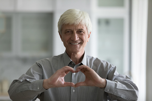 Cheerful grey haired old man showing hand heart at chest, looking at camera with happy toothy smile, expressing romance, love, happiness, gratitude. Senior male blogger showing like shape