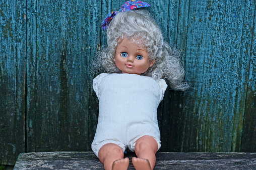 one old brown plastic doll without hands in a white dress sits on a gray table against a green wooden wall