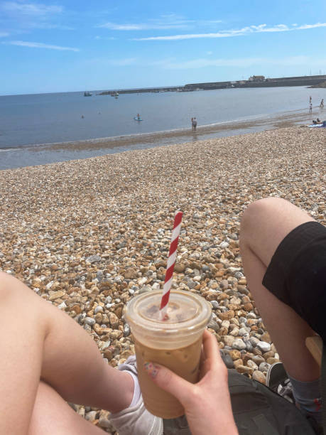Stock photo showing an unrecognisable woman holding a plastic, disposable cup of iced latte coffee whilst sat besides unrecognisable man on the shingle beach overlooking the Cobb and harbour at the seaside town of Lyme Regis, Dorset, England, UK.
