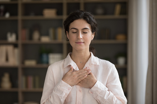 Portrait of young attractive woman standing alone in living room put their folded crossed palms on chest feel grateful, express appreciation, sincere feelings with eyes closed. Belief, charity concept