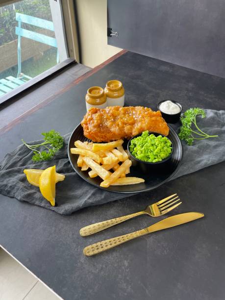 Stock photo showing the set up of a food photography studio photo shoot. imagen de la sesión de fotos del estudio de fotografía de alimentos detrás de escena configurada, tomando fotos en el interior de pescado, papas fritas y guisantes blandos en plato negro, guarnición de perejil, salsa tártara, cuchillo y tenedor, juego