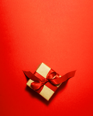 Brown paper gift box with a red satin ribbon bow on top on red background. Christmas holiday mother and father Valentines days and birthday present flat lay concept with copy space.