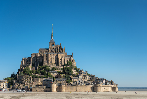Mont St. Michel, Normandy, France - July 8, 2022: The brown-stone structure on its rock surrounded by sandy tidal flats against blue sky. Some delivery vans at main entrance.