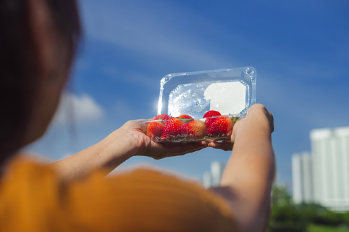 Human hands carrying fresh, juicy and ripe strawberries placed in a plastic container isolated on city sky background.