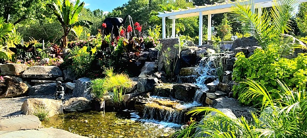 Sunny summer day showing a large waterfall amidst lush plants and foliage and a white gazebo in the background.