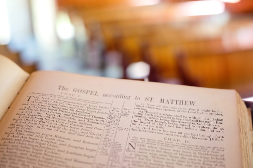 Old Bible from the 1880s open to the beginning of the Gospel of Matthew with church pews in the distance.