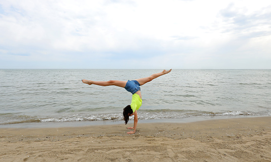 slender teenage girl performs a split with long legs on the seashore in summer
