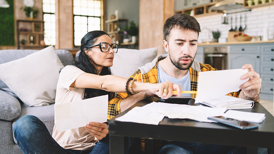 Couple calculating bills at home using calculator.  Couple checking bills and documents while calculating finances sitting in the living room. Man and woman at home analyzing their finance with documents.