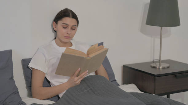 istock Hispanic Woman Reading Book while Sitting in Bed 1423254658
