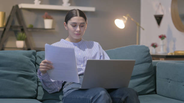 istock Hispanic Woman with Laptop Working on Documents on Sofa 1423257842