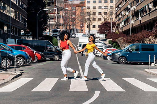 Pretty african american young women holding hands and walking in zebra crossing in the city and having fun.