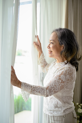 Portrait, A happy Asian middle-aged woman looking out the window in the morning, taking in the beautiful view outside the window.