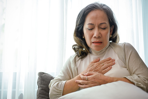 A sick Asian middle-aged woman with heart disease or heart ache, touching her chest and having a heart attack while relaxing at home. Health care concept