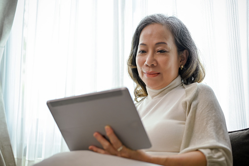 A beautiful Asian-aged woman watches a video clip online on a digital tablet touchpad in her comfortable living room.