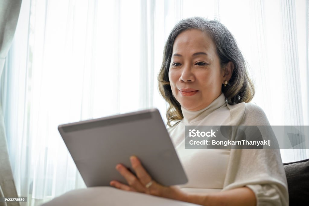 A beautiful Asian-aged woman watches a video clip online on a digital tablet in her living room. A beautiful Asian-aged woman watches a video clip online on a digital tablet touchpad in her comfortable living room. Computer Stock Photo