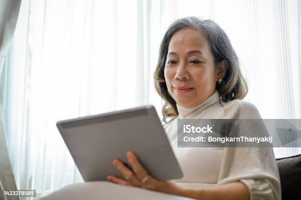 A Beautiful Asianaged Woman Watches A Video Clip Online On A Digital Tablet In Her Living Room Stock Photo - Download Image Now