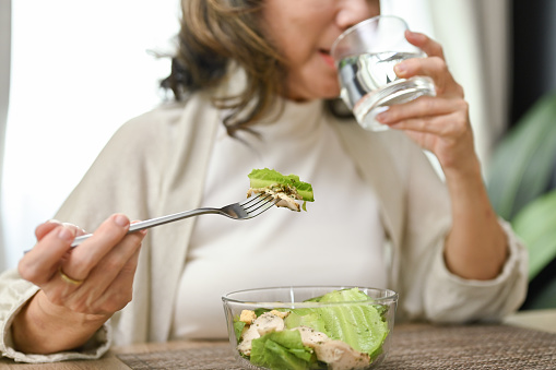 Healthy and happy Asian-aged eating salad bowl and drinking water in her dining room. cropped and close-up image
