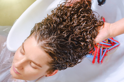 Professional hairstylist washes client’s hair with conditioner shampoo for washing. Close up of curlers in hair. Creating hairstyles by a hairdresser in the salon. Shallow depth of field