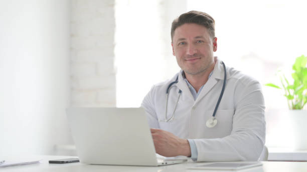 istock Male Doctor Smiling at Camera while using Laptop in Office 1423284907