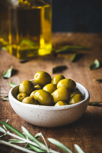 A bowl full of green olives, with an olive oil bottle behind, on a rustic wooden table