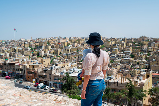 Woman in a baby in a baby carrier visiting Amman in Jordan overlooking the old city cityscape on a sunny day. Traveler in Amman historical area. Young mother traveling abstract with copy space